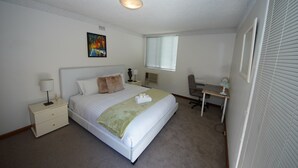 A bedroom with a bed, bedside table, lamp, desk, chair, and a window.