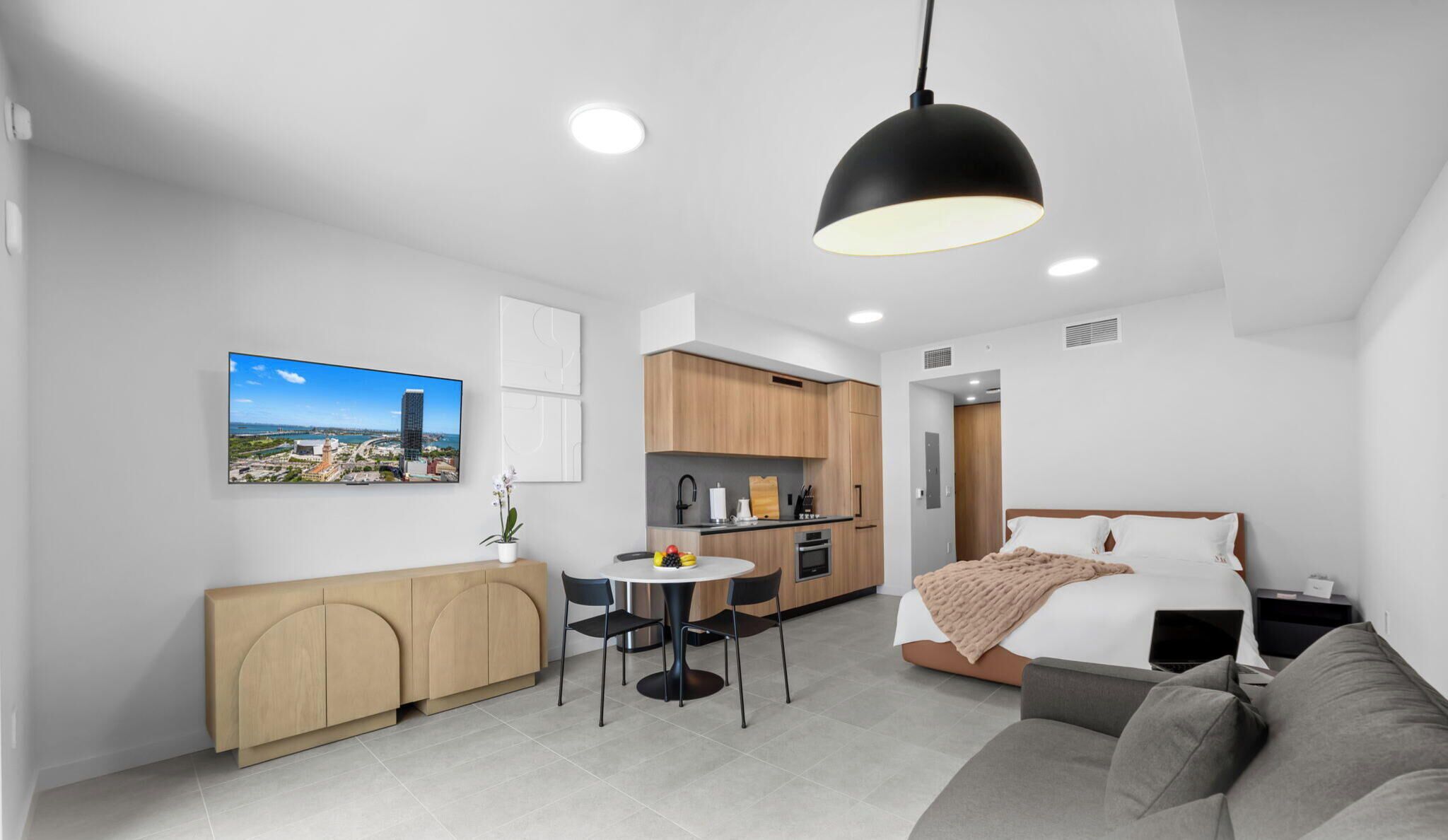 Ein modernes Hotelzimmer mit Bett, Sofa, Esstisch mit Stühlen, Küchenzeile und Wandbild mit Stadtblick.