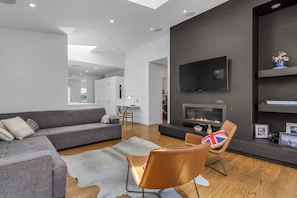Ein modernes Wohnzimmer mit grauem Sofa, Ledersessel, Flachbildfernseher, Kamin und einem Essbereich im Hintergrund.