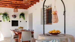 Zona de menjador amb taula de fusta, cadires, plantes penjants i un plat de fruita.