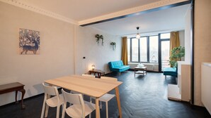 Um quarto com uma mesa de jantar de madeira, cadeiras brancas, um sofá azul e uma janela com cortinas.
