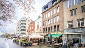 A street with outdoor seating, a building labeled 'APALACE,' and another with a sign 'STELLA ARTOIS.' - The Industrial Apartments|2BBK|NS (Antwerp)