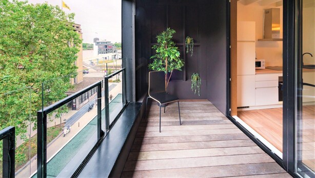 A balcony with a wooden floor, a chair, a potted plant, and a view of a city street. - The Industrial Apartments|2BBK|NS (Antwerp)