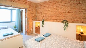 A room with a brick wall, two beds with patterned bedding, a small plant, and a window with curtains. - The Industrial Apartments|2BBK|NS (Antwerp)