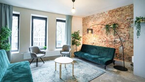 A living room with a brick wall, two windows, a green sofa, a round coffee table, and two chairs. - The Industrial Apartments|2BBK|wifi|NS (Antwerp)