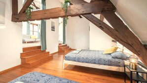 Ein Schlafzimmer im Loft-Stil mit freiliegenden Holzbalken, einem Bett mit gemusterten Textilien, einem Nachttisch mit Lampe und einer Treppe zu einer oberen Ebene.