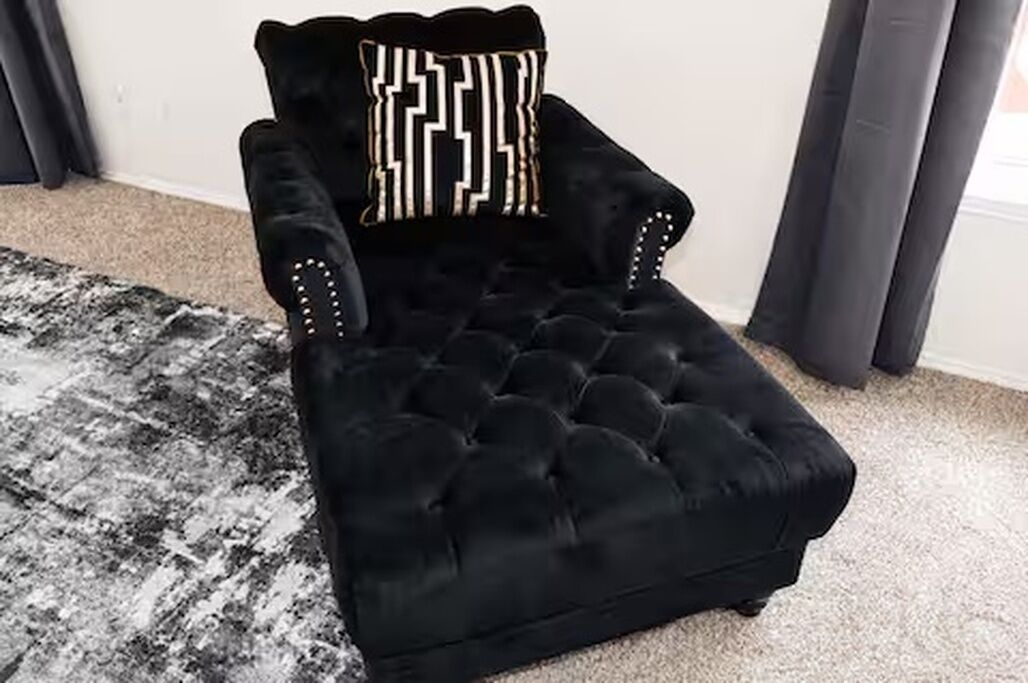 Een zwarte fauteuil met een versierde kussen, geplaatst op een kamerkleed in een kamer met een effen muur en gordijnen.