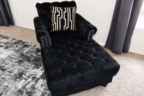 Un fauteuil noir à accoudoirs capitonnés, agrémenté d’un coussin décoratif, est placé sur un tapis à motifs dans une pièce dont le mur est uni et qui est dotée de rideaux.