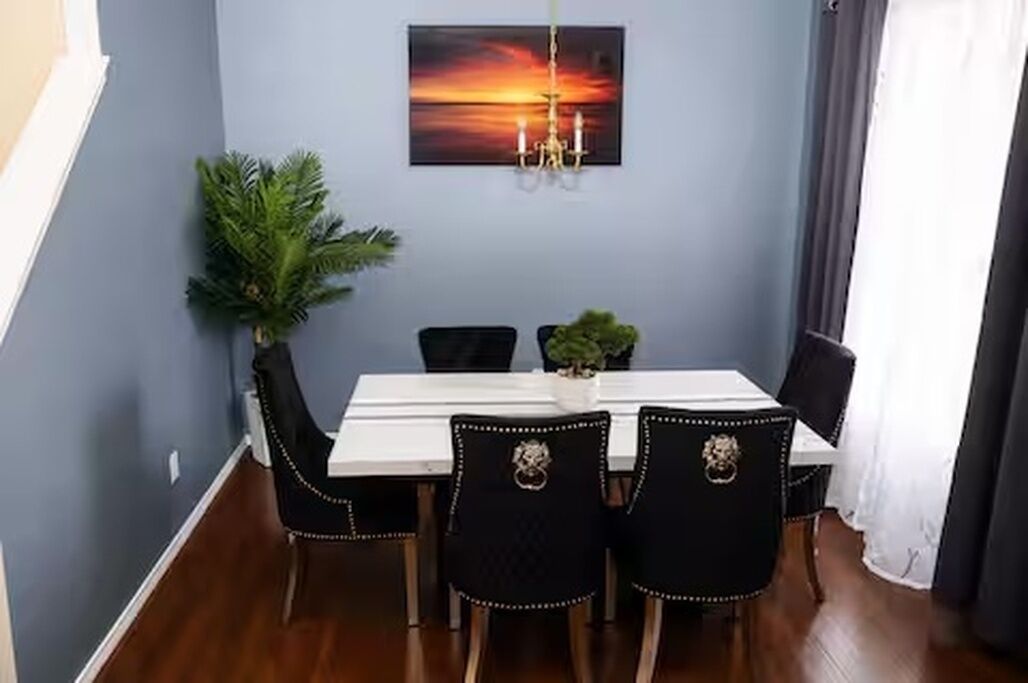 Ein Essbereich mit einem weißen Tisch, schwarzen Stühlen, einer Kristallleuchtre und einem Gemälde von einem Sonnenuntergang an der Wand.