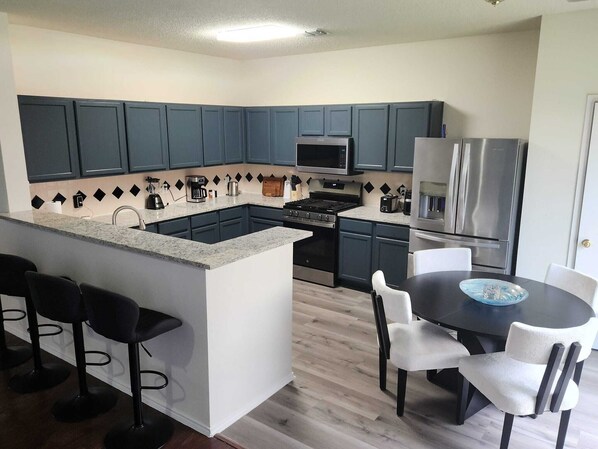 Una cocina moderna con armarios azul oscuro, una isla central con taburetes de bar, una mesa de comedor con sillas y un refrigerador de acero inoxidable.