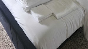 Una cama bien hecha con ropa de cama blanca y toallas dobladas, un cabecero oscuro y un sillón gris en una habitación de hotel.
