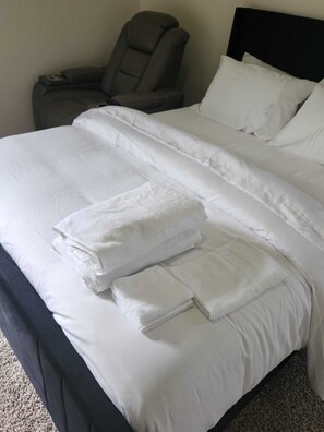 Una cama bien hecha con ropa de cama blanca y toallas dobladas, un cabecero oscuro y un sillón gris en una habitación de hotel.