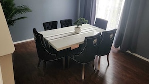 Un comedor con una mesa blanca, sillas tapizadas de color negro, una araña, una planta en maceta y un cuadro en la pared.