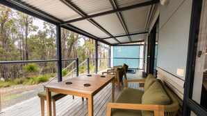 A covered outdoor seating area with a wooden table and chairs, a sofa, and a view of the surrounding forest.