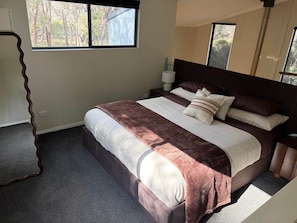 A bedroom with a large bed, a bedside table, a lamp, and a view of trees through the window.