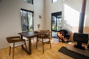 Ein modernes Interieur mit einem Holzesstisch, Stühlen, einem Kamin und großen Fenstern mit Blick ins Freie.
