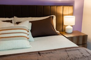 A neatly made bed with a striped pillow, a solid brown pillow, and a white pillow. There is a bedside table with a lamp and a nightstand.