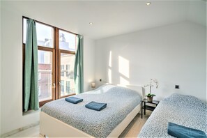 Une chambre moderne avec une grande fenêtre, un lit recouvert d’une couvre-lit à motifs, une table de chevet avec une lampe et une petite plante en pot.
