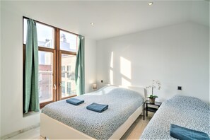 Quarto moderno com uma janela grande, uma cama com colcha estampada, mesa de cabeceira com abajur e uma pequena planta em vaso.