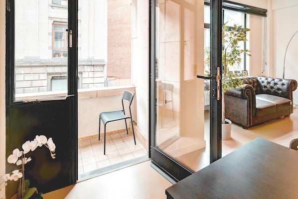 En moderne stue med sofa, spisebord, stol og en balkon med udsigt til bygninger.