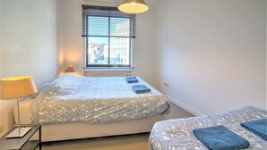 A bedroom with a bed, a bedside table with a lamp, a window with blinds, and a view of buildings outside. - Welcoming Antwerp Relaxing Stay (Antwerp Center)