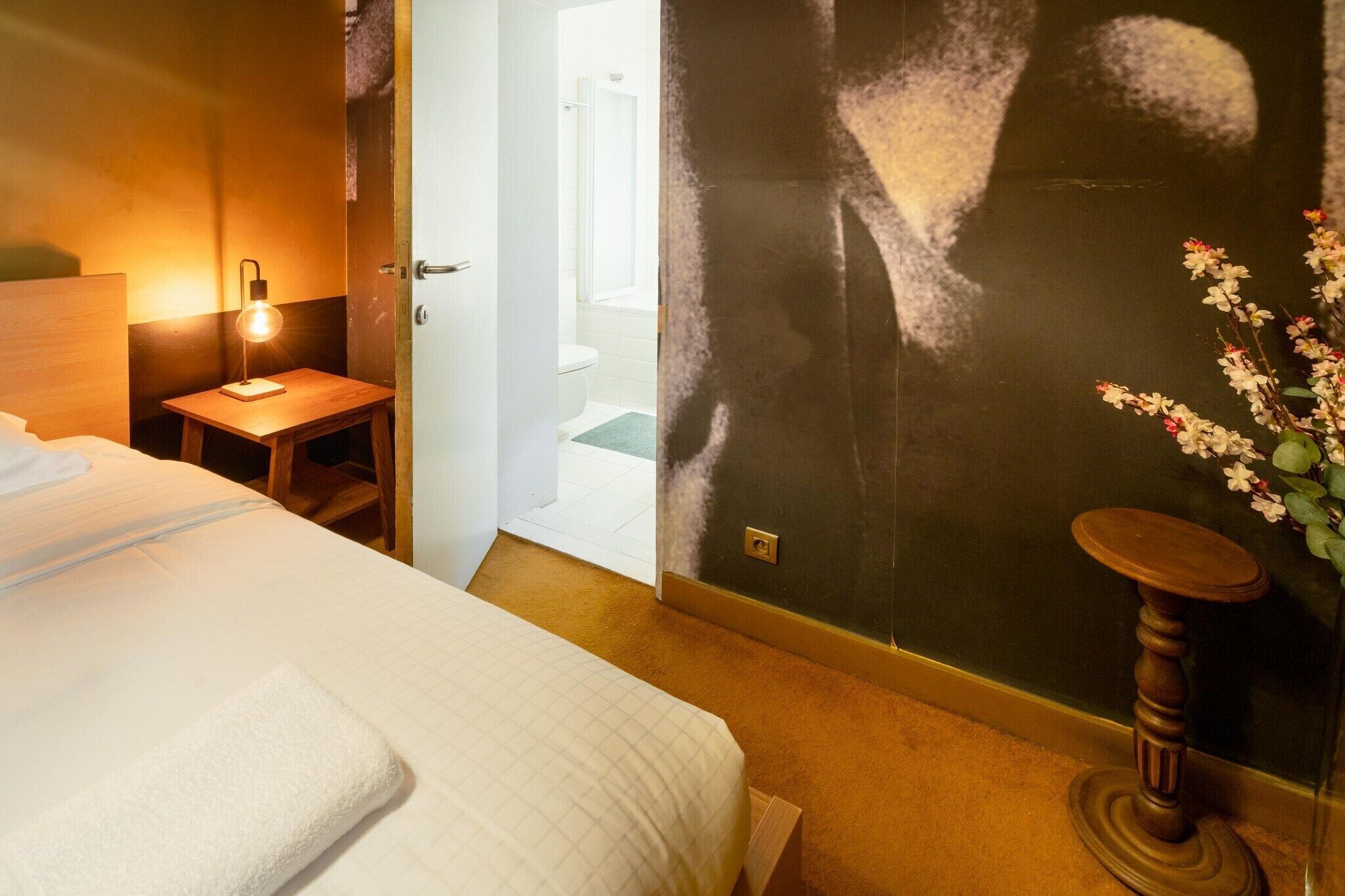 A hotel room with a bed, a bedside table with a lamp, a small stool, a vase with flowers, and a door leading to a bathroom.