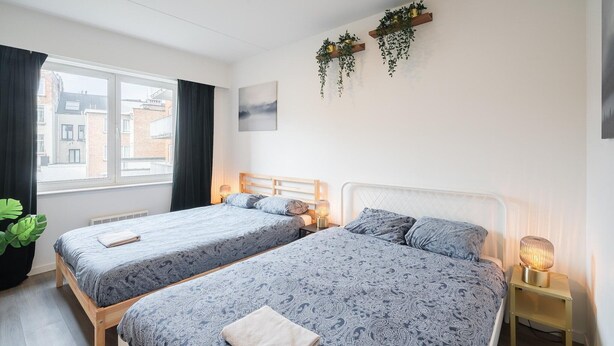 A hotel room with two beds, a window with curtains, a radiator, a wall-mounted plant, and a bedside table with a lamp. - Antwerp bright apartment stay (Antwerp)