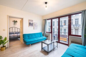 A room with a blue sofa, a glass coffee table, a bed with a metal frame, a window with a view of buildings, and a plant. - Stay in the heart of Antwerp (Antwerp)