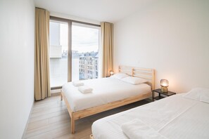 Et soveværelse med et stort vindue, en seng, et natbord med lampe, og en anden seng.