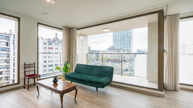 Un salon spacieux avec un canapé vert, une chaise en bois, une petite table et une plante en pot. De grandes fenêtres offrent une vue sur le paysage urbain.