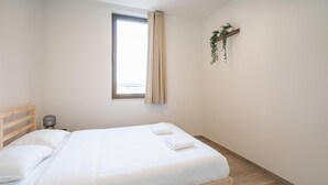 Een net opgemaakt bed met witte lakens, een houten hoofdbord, een bedlampje en een raam met gordijnen.