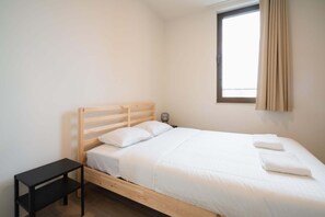 Un lit simple avec une tête de lit en bois, une table de chevet, une lampe et une fenêtre avec des rideaux.