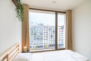 Ett hotellrum med ett stort fönster, en säng, ett sängbord med en lampa och en hängande växt.