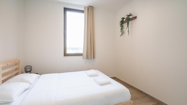 A neatly made bed with white linens, a wooden headboard, a bedside lamp, and a window with curtains. - Spacious City Retreat with Balcony (Antwerp)