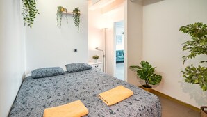 Un dormitorio con una cama, dos almohadas, una mesita de noche con una planta y vistas a otra habitación.