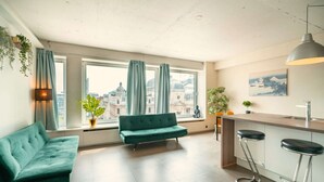 Un apartamento moderno con sofá, un pequeño rincón de descanso, una cocina con taburete de bar y vistas a edificios a través de amplios ventanales.