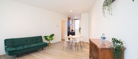 A modern living room with a green sofa, a dining table with chairs, a wooden dresser, and a kitchen in the background.