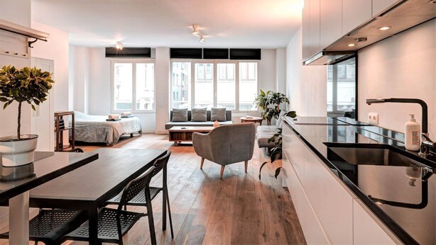 Una cocina moderna con cubierta negra, una zona de comedor con mesa y sillas de madera, un sofá y un ventanal con vistas a edificios.