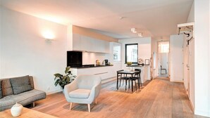 Una sala moderna con sofá, sillón y mesa de centro, una cocina con armarios blancos y televisor, y un comedor con mesa y sillas.