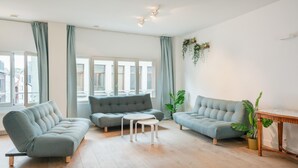 Una sala moderna con un sofá gris, un sillón gris más pequeño y una mesa de centro blanca. Hay ventanas grandes con cortinas, piso de madera y una planta a la derecha.