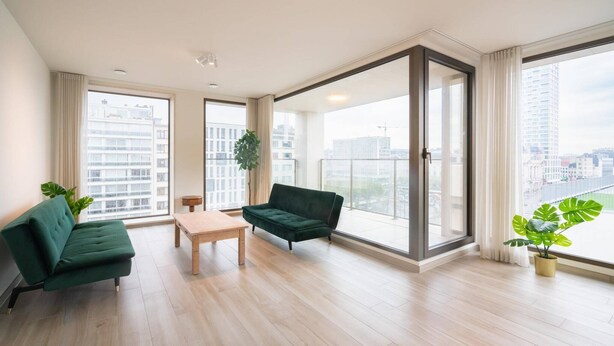 Un salon spacieux avec un canapé vert, une table basse en bois et de grandes fenêtres donnant sur la ville.