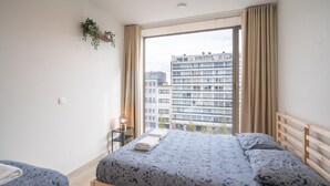 Une chambre avec une grande fenêtre, un lit recouvert d’une literie à motifs, une table de chevet avec une lampe, et une vue sur un bâtiment à l’extérieur.