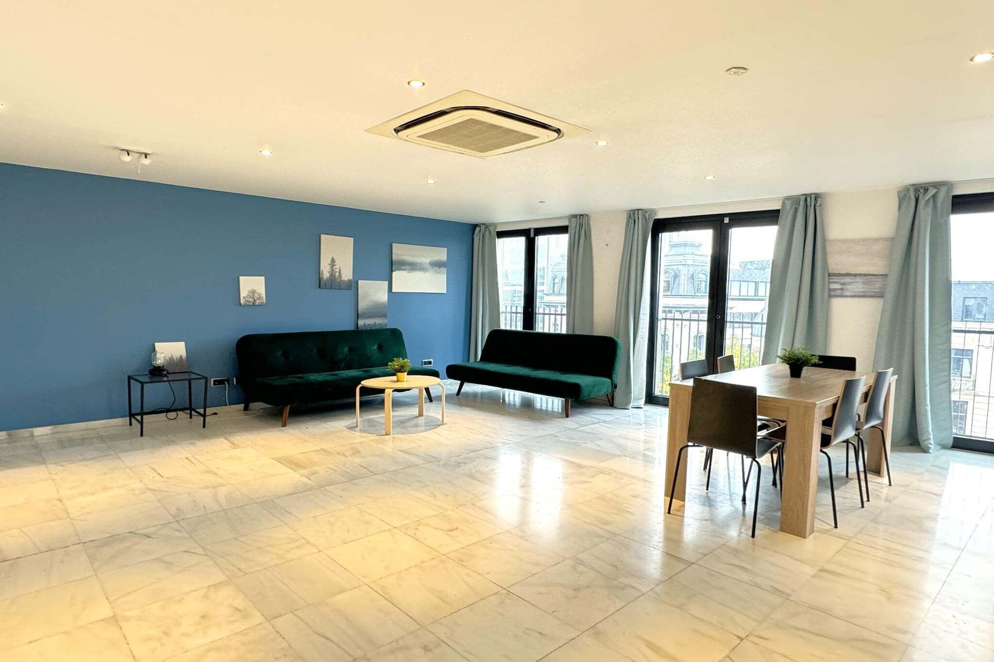 Un salon moderne avec un mur bleu, un canapé vert, une petite table basse, une table à manger avec des chaises et de grandes fenêtres avec des rideaux.