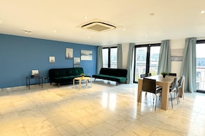 Een moderne woonkamer met een blauwe wand, een groene bank, een klein bijzettafeltje, een eettafel met stoelen en grote ramen met gordijnen.