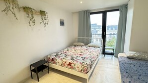 Une chambre avec un lit, une petite table, un balcon avec vue et des plantes suspendues.