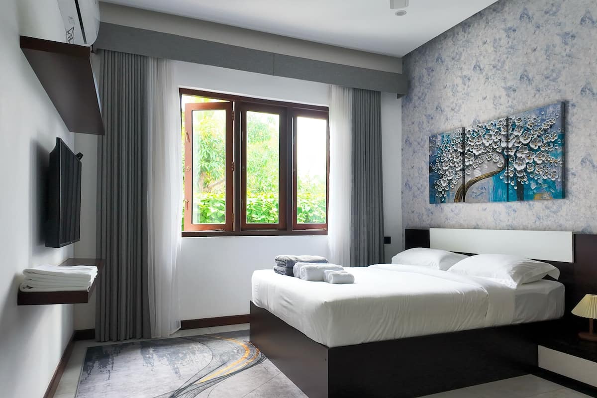 A bedroom with a bed, a painting, a window, and a bedside table.