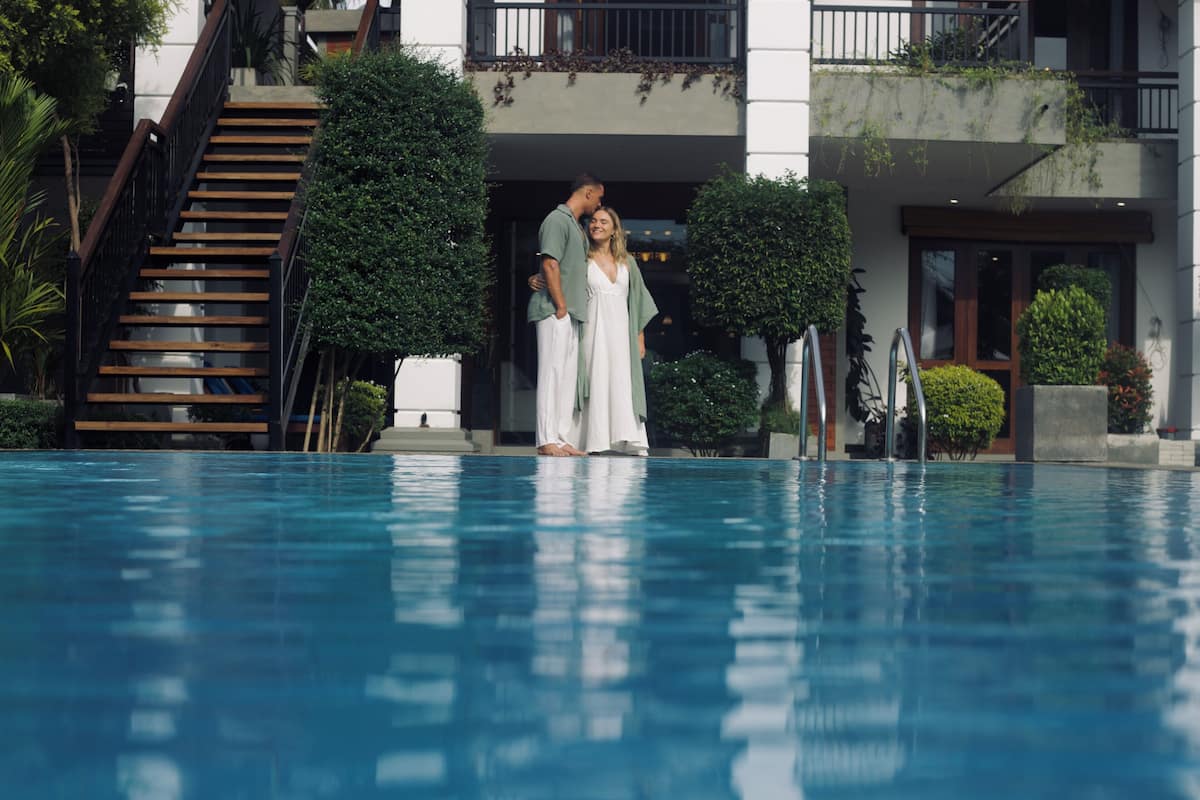 A swimming pool with a wooden staircase and a building with balconies and greenery in the background.