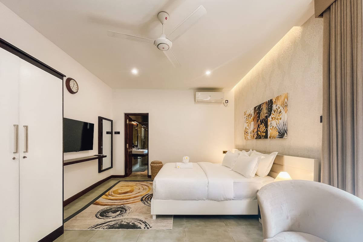 A hotel room with a bed, a sofa, a TV, a mirror, a ceiling fan, and a bathroom visible through an open door.
