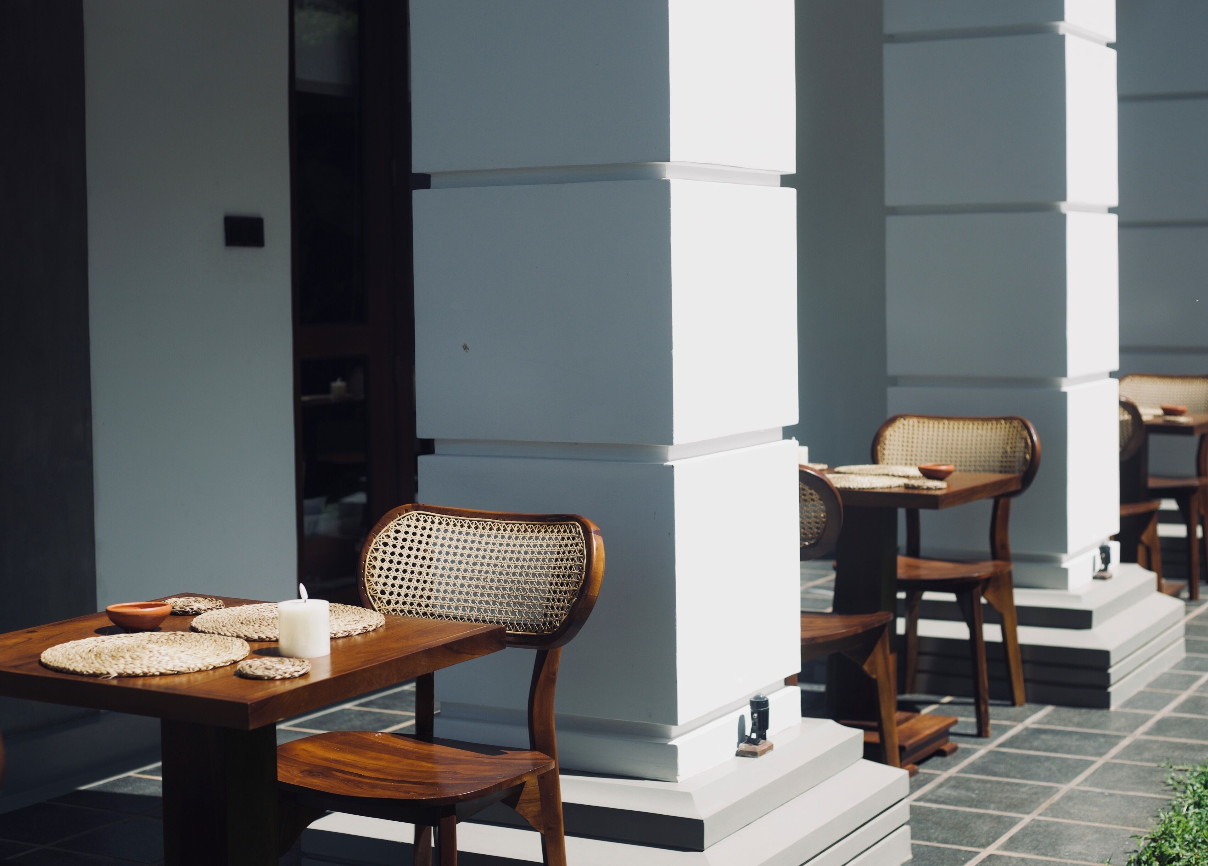 A row of wooden tables and chairs with woven mats, set against white pillars on a tiled floor.