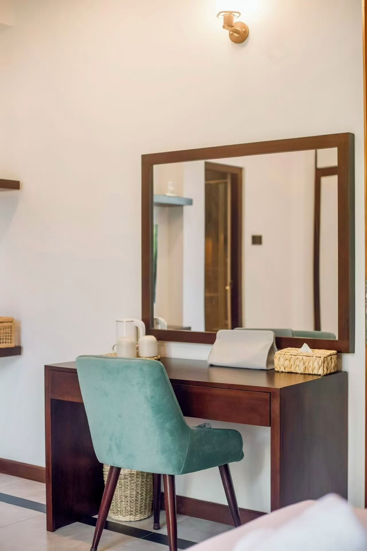 A hotel room with a wooden desk, a teal chair, a large mirror, and a bedside table with a lamp and tissues.